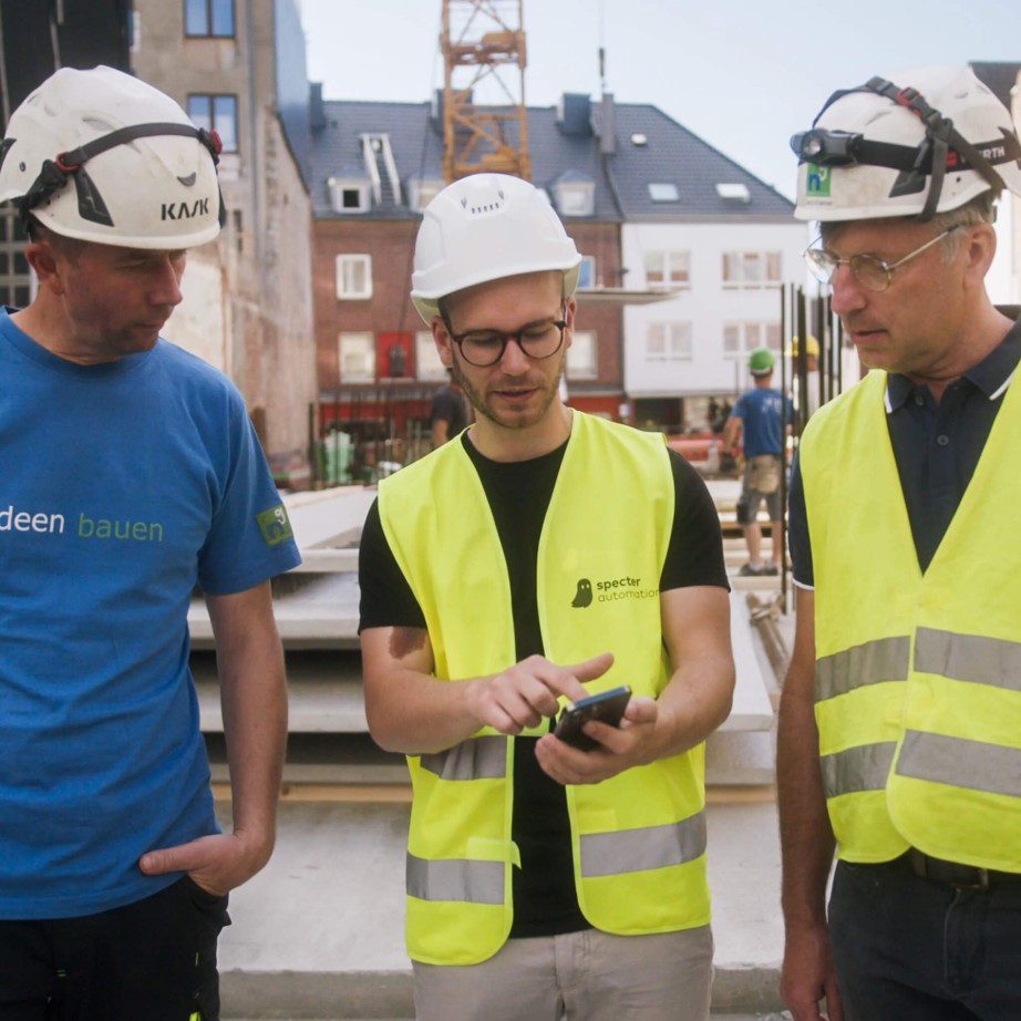 specter Mitarbeiter zeigt die Bausoftware Vorteile am Smartphone für einen Polier und einen Bauleiter. Im Hintergrund ist die Baustelle zu sehen.